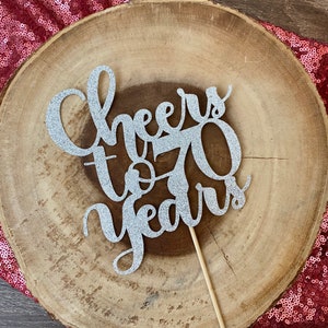 Puede incluir: Topper de pastel de purpurina plateada con el texto "Cheers to 70 Years" sobre un fondo de madera.