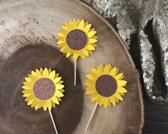 May include: Three yellow paper sunflower cupcake toppers with brown glitter centers on a wooden surface.