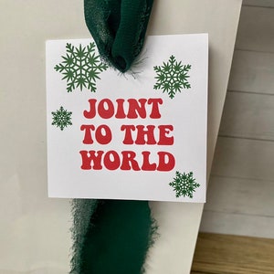 May include: White gift tag with green snowflakes and red text that reads "Join to the World". The tag is tied with a green ribbon.
