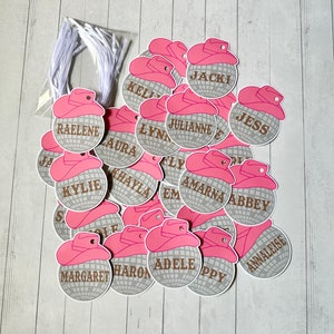 May include: A pile of silver and pink party favor tags with names printed on them. The tags are shaped like disco balls with a pink cowboy hat on top.