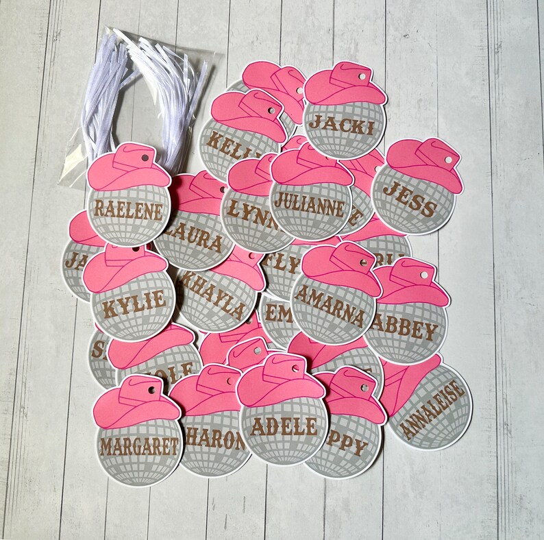May include: A pile of silver and pink party favor tags with names printed on them. The tags are shaped like disco balls with a pink cowboy hat on top.