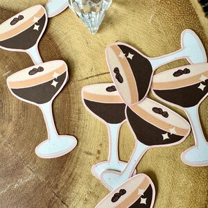 May include: Paper cutouts of espresso martini glasses with brown, tan, and white colors. Each glass features a coffee bean and star accents. The glasses are arranged on a wooden surface, suggesting a cocktail-themed craft or decoration.