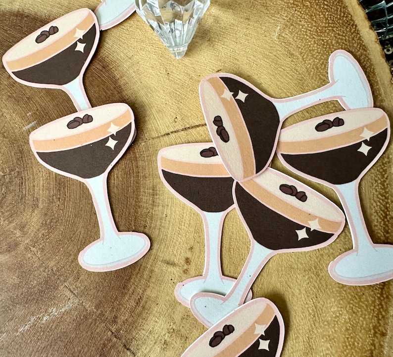 May include: Paper cutouts of espresso martini glasses with brown, tan, and white colors. Each glass features a coffee bean and star accents. The glasses are arranged on a wooden surface, suggesting a cocktail-themed craft or decoration.