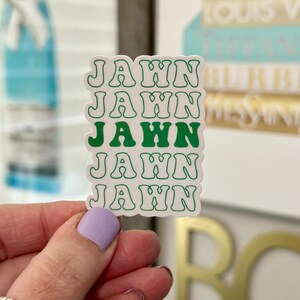 May include: A white sticker with the word "JAWN" stacked five times in a green, retro font. The sticker is held by a hand with a light purple painted nail. The background includes blurred images of books and gold lettering.
