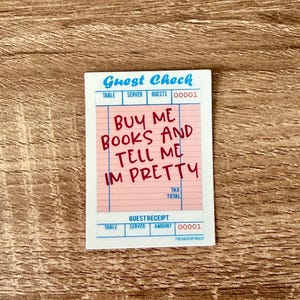 May include: A sticker resembling a guest check, featuring the text "BUY ME BOOKS AND TELL ME IM PRETTY" in red handwritten font. The top of the sticker reads "Guest Check" in blue, with fields for table, server, and guests.