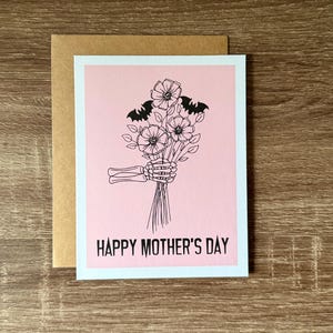 Puede incluir: Una tarjeta de felicitaci&oacute;n con un fondo rosa y una ilustraci&oacute;n en negro de una mano de esqueleto sosteniendo un ramo de flores y dos murci&eacute;lagos. Las palabras "HAPPY MOTHER'S DAY" est&aacute;n impresas en negro en la parte inferior de la tarjeta. La tarjeta va acompa&ntilde;ada de un sobre marr&oacute;n.