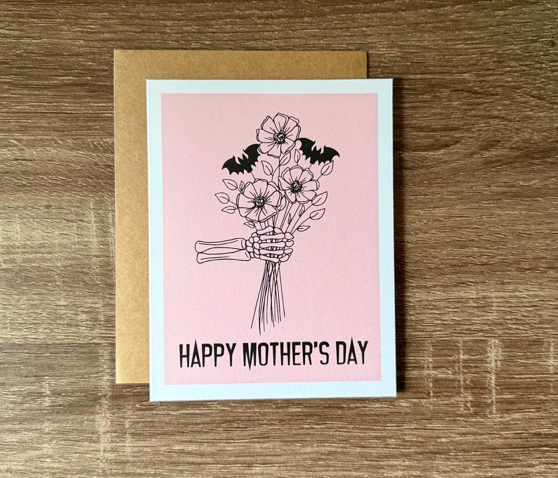 Puede incluir: Una tarjeta de felicitaci&oacute;n con un fondo rosa y una ilustraci&oacute;n en negro de una mano de esqueleto sosteniendo un ramo de flores y dos murci&eacute;lagos. Las palabras "HAPPY MOTHER'S DAY" est&aacute;n impresas en negro en la parte inferior de la tarjeta. La tarjeta va acompa&ntilde;ada de un sobre marr&oacute;n.