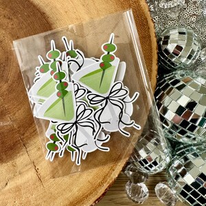 Puede incluir: Una bolsa de pl&aacute;stico transparente llena de pegatinas de vaso de martini verde y rojo. Las pegatinas tienen una aceituna verde y una cereza roja en un palillo.