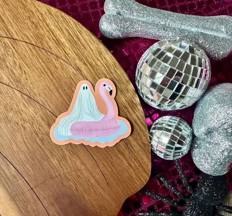 May include: A sticker featuring a ghost and a pink flamingo pool float. The ghost is draped over the flamingo, floating on a blue and orange water background. The sticker is on a wooden surface, with disco balls and a glittery bone in the background.