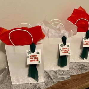 May include: Three white gift bags with green and red ribbon and tags. The tags have the text "Joint to the World", "Santa's Favorite Stoner", and "Merry Kushmas".