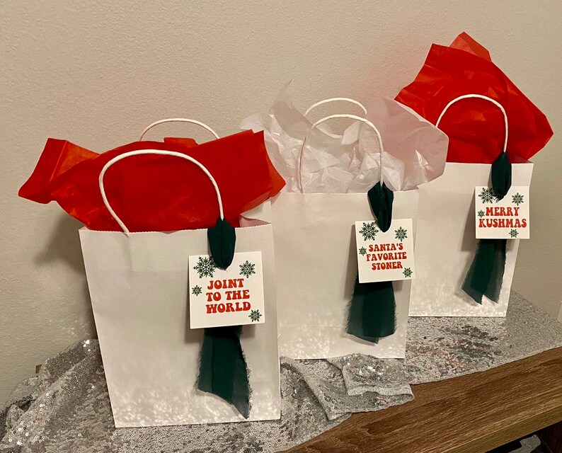 May include: Three white gift bags with green and red ribbon and tags. The tags have the text "Joint to the World", "Santa's Favorite Stoner", and "Merry Kushmas".