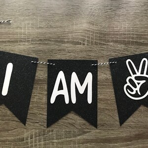 May include: Black and white banner with the words "I AM" and a peace sign. The banner is made of felt and has a glitter finish.