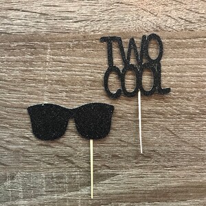 Könnte beinhalten: Zwei schwarze Glitzer-Kuchentopper, einer sagt "Two Cool" und der andere ist eine Sonnenbrille.