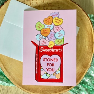 May include: A pink card with a red box that says "Sweethearts" and "Stoned for You" in a heart shape. The box is overflowing with colorful candy hearts that have phrases like "Love Stoned", "High Love You", "Let's Smoke", and "Hug Me".