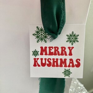 May include: White gift bag with a green ribbon and a gift tag that says "Merry Kushmas" in red letters with green snowflakes.