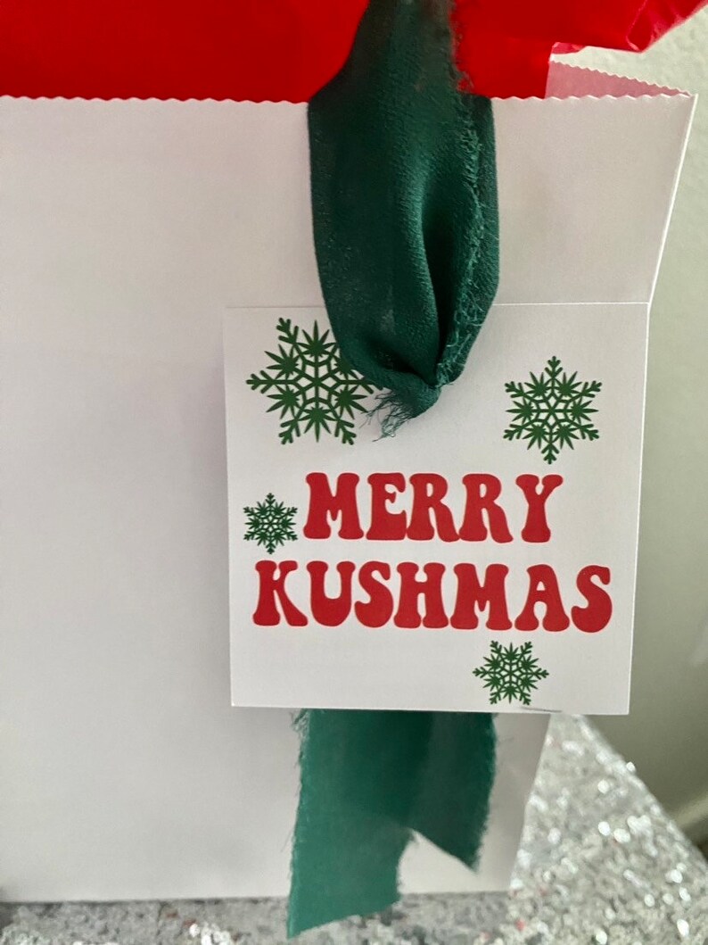 May include: White gift bag with a green ribbon and a gift tag that says "Merry Kushmas" in red letters with green snowflakes.