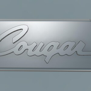 Könnte beinhalten: Ein silberfarbenes, ovalförmiges Metallschild mit dem Wort "Cougar" in einer stilisierten Schrift. Das Schild hat ein kleines Loch auf der linken Seite und einen abgerundeten Rand. Der Hintergrund ist einfarbig hellgrau.