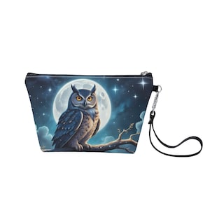 May include: A cosmetic bag featuring a detailed illustration of an owl perched on a branch under a full moon and starry night sky. The bag has a black zipper and a black wrist strap. The bag is a trapezoid shape.