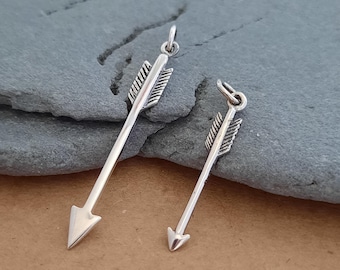 May include: Two silver arrow charms, one larger than the other, with a detailed arrowhead and fletching. The charms are displayed on a gray stone surface.