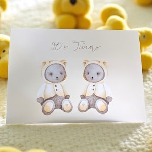 May include: A greeting card with the text "It's Twins" features watercolor illustrations of two teddy bears wearing hooded jackets. The bears are gray with tan accents, set against a soft, off-white background. The card is surrounded by yellow plush toys.