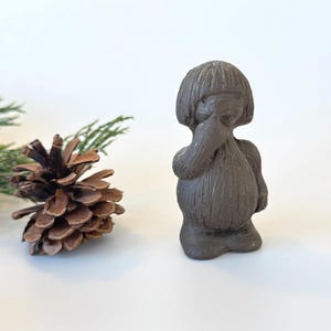May include: A small, dark brown ceramic figurine of a person with a hand raised to their face. A pine cone and a sprig of evergreen are to the left of the figurine, set against a white background.