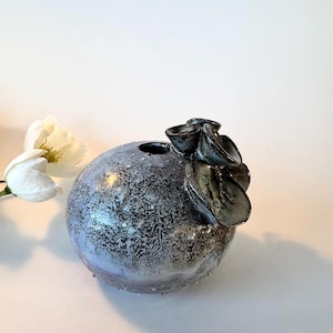 Op de afbeelding: Een ronde, gespikkelde keramische vaas met een donkergrijze en zilveren afwerking. De vaas heeft een cluster van donkere, bladachtige vormen aan de bovenkant. Een witte bloem staat op de achtergrond.