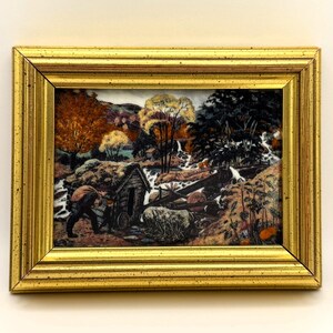 Op de afbeelding: Een ingelijst schilderij dat een landelijk tafereel voorstelt met een klein houten huisje, een beekje en figuren die in het bos werken. Het schilderij speelt zich af in de herfst met kleurrijk gebladerte in tinten bruin, oranje en geel.