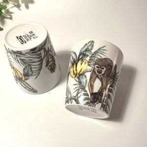 Puede incluir: Dos tazas de cerámica blanca con un diseño de jungla. Una taza está en posición vertical, la otra está inclinada. El diseño presenta un mono, plátanos y hojas tropicales en tonos verdes, amarillos y marrones.