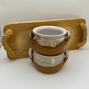 May include: Two brown and white ceramic mugs with handles stacked on top of each other on a wooden tray with rope handles.