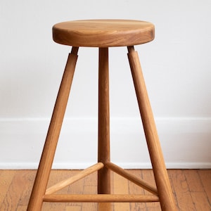 May include: A wooden stool with a round seat and three legs. The stool is made of natural wood and has a simple, rustic design.