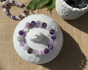 May include: A purple amethyst and clear quartz beaded bracelet with silver accents. The bracelet is on a white ceramic dish with a floral design.