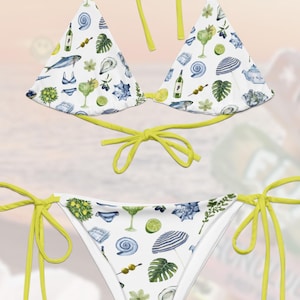 May include: A white bikini with a yellow trim and a tropical print featuring cocktails, limes, and seashells. The bikini top is a triangle style and the bottom is a cheeky style with ties on the sides.