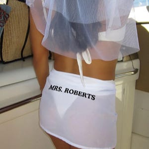 May include: A bride-to-be wears a white veil and a sheer white skirt. The skirt has the text "MRS. ROBERTS" printed in black. The bride is on a boat, with a glimpse of the water and sky in the background.