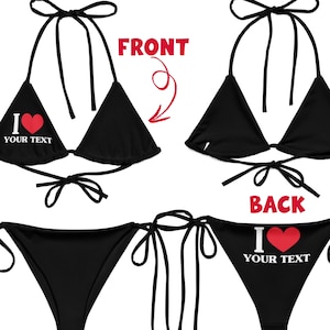Può includere: Set bikini nero con chiusure a fiocco. La parte anteriore del top e la parte posteriore del fondo presentano la scritta "I ❤️ YOUR TEXT" con un cuore rosso. L'immagine mostra anche il retro del top del bikini e il davanti del fondo.
