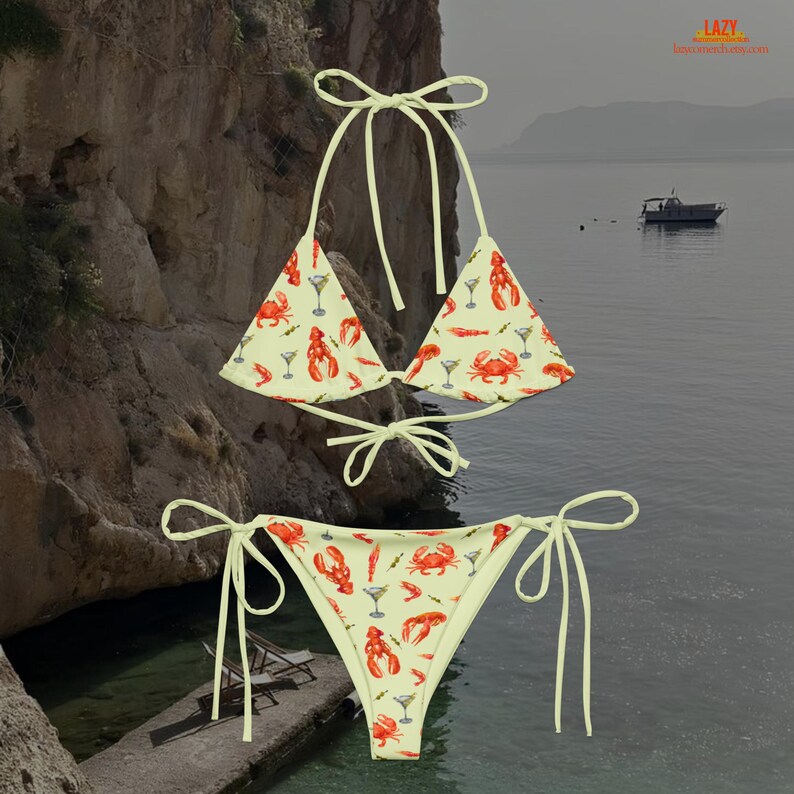 K&ouml;nnte beinhalten: Ein zweiteiliges Bikini-Set mit hellgelbem Hintergrund und einem Muster aus roten Hummern, Krabben, Garnelen und Martinigl&auml;sern. Das Bikini-Oberteil ist im Triangel-Stil mit Bindetr&auml;gern, und das Unterteil ist ein klassischer Schnitt mit Bindeb&auml;ndern.