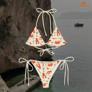 K&ouml;nnte beinhalten: Zweiteiliger Bikini mit wei&szlig;em Hintergrund und einem sich wiederholenden Muster aus roten Hummern, Krabben und Martinigl&auml;sern. Das Oberteil ist im Triangel-Stil mit wei&szlig;en Bindeb&auml;ndern.