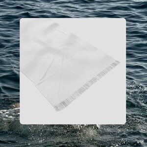 May include: A white rectangular scarf with a fringed edge. The scarf is made of a lightweight fabric and appears to be folded. The background is a blurred image of water.