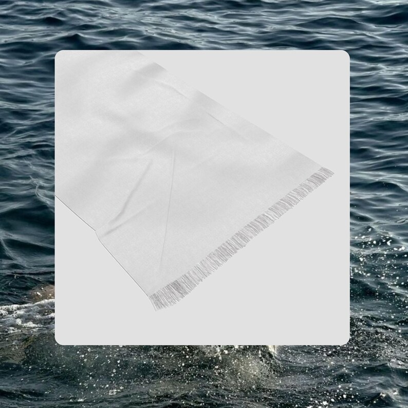 May include: A white rectangular scarf with a fringed edge. The scarf is made of a lightweight fabric and appears to be folded. The background is a blurred image of water.