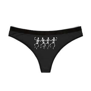 Puede incluir: Bragas negras con un gráfico blanco de tres esqueletos bailando. La ropa interior tiene una cinturilla negra y una parte trasera tipo tanga. Los esqueletos están representados en un estilo juguetón y caricaturesco.