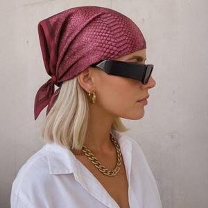 May include: A woman with short blonde hair is wearing a white button-down shirt, a gold chain necklace, and a burgundy bandana with a snake skin pattern. She is also wearing black sunglasses and gold hoop earrings.