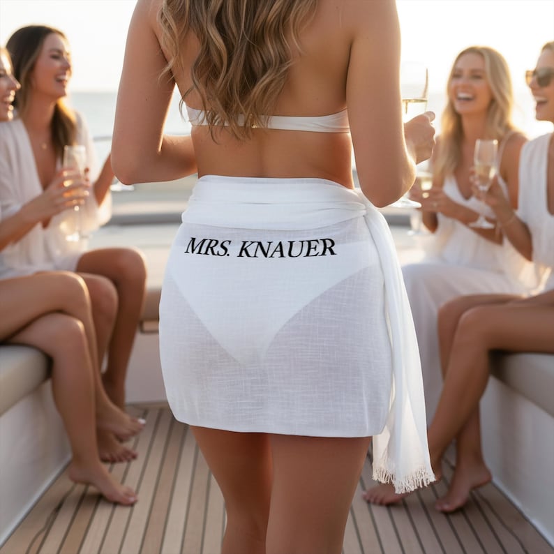 May include: A white sarong with the text "MRS. KNAUER" in black, worn over a white bikini bottom. Several women are in the background, some holding champagne flutes. The scene takes place on a boat.