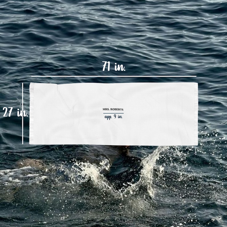 May include: A white beach towel, 71 inches long and 27 inches wide, floats on water. The towel has the text "MRS. ROBERTS" and "app. 9 in." printed on it. The water is dark blue with white splashes.
