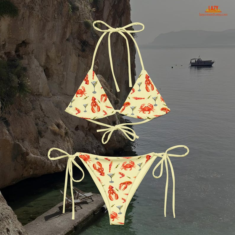 K&ouml;nnte beinhalten: Zweiteiliger Bikini mit cremefarbenem Hintergrund und einem sich wiederholenden Muster aus roten Hummern, Krabben und Martinigl&auml;sern. Das Bikinioberteil ist im Dreiecksstil mit Bindeb&auml;ndern und das Unterteil im klassischen String-Stil.