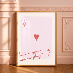 May include: A framed print featuring a pink playing card design. The card shows the Ace of Hearts with a red heart symbol and the text "Love's a game, wanna play?" in a cursive font. The frame is gold.