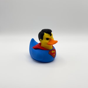 May include: A rubber ducky in the style of Superman. The duck has a yellow head with black hair and an orange beak. It wears a blue outfit with a red cape and the Superman logo on the chest.