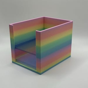 May include: A rainbow-colored desk organizer with two shelves. The organizer is a cube shape with a pastel color gradient, transitioning from pink to blue. The shelves are transparent, and the overall design is modern and minimalist.
