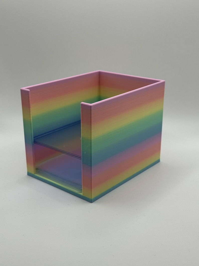 May include: A rainbow-colored desk organizer with two shelves. The organizer is a cube shape with a pastel color gradient, transitioning from pink to blue. The shelves are transparent, and the overall design is modern and minimalist.