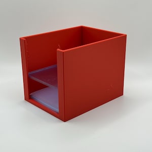 May include: A bright red, rectangular desk organizer with two clear plastic shelves. The organizer is made of solid material and has a simple, modern design. The shelves are designed to hold small items.