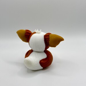 Może przedstawiać: Wydrukowana w 3D figurka Gremlina, z białym korpusem i brązowymi i beżowymi akcentami. Figurka ma duże, spiczaste uszy i małą, kolczastą koronę. Figurka jest w pozycji siedzącej.