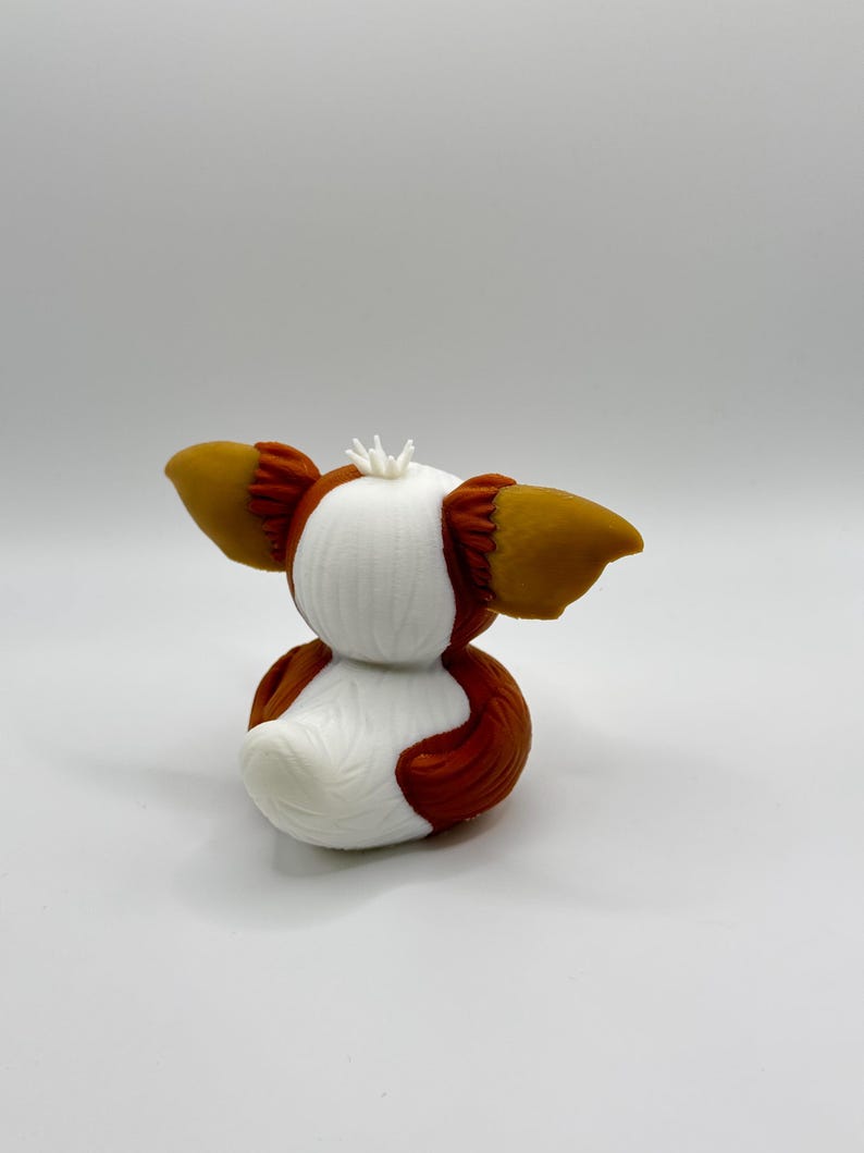 Może przedstawiać: Wydrukowana w 3D figurka Gremlina, z białym korpusem i brązowymi i beżowymi akcentami. Figurka ma duże, spiczaste uszy i małą, kolczastą koronę. Figurka jest w pozycji siedzącej.